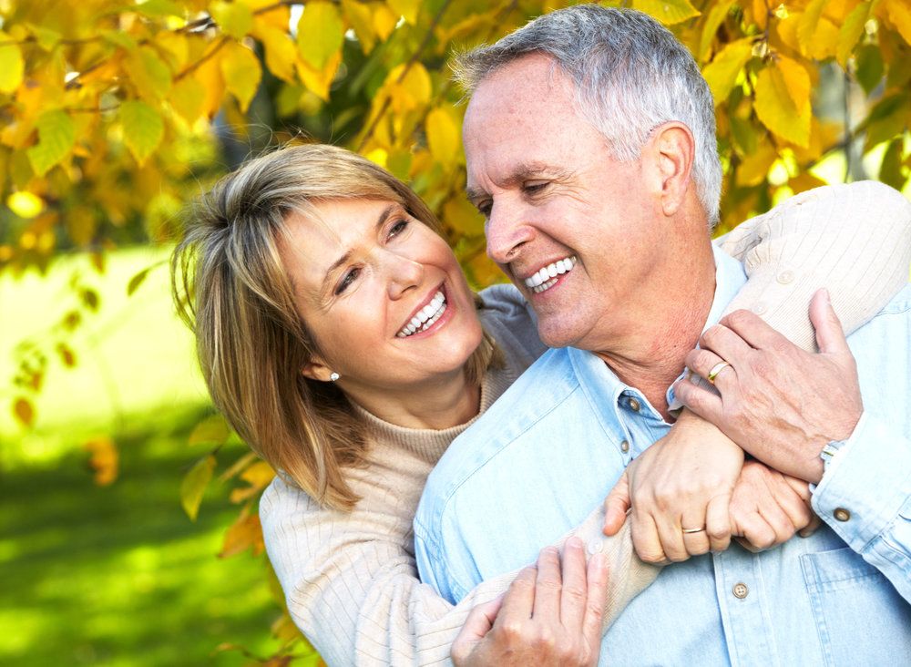 A middle-aged woman hugs a man from behind while the two stand in front of a brightly colored tree.